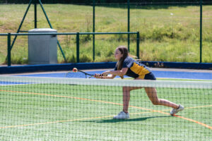 A pupil, wearing a blue and yellow P.E kit, it playing tennis on an outdoor tennis court. She is reaching over to hit the ball with the racket in her right hand.