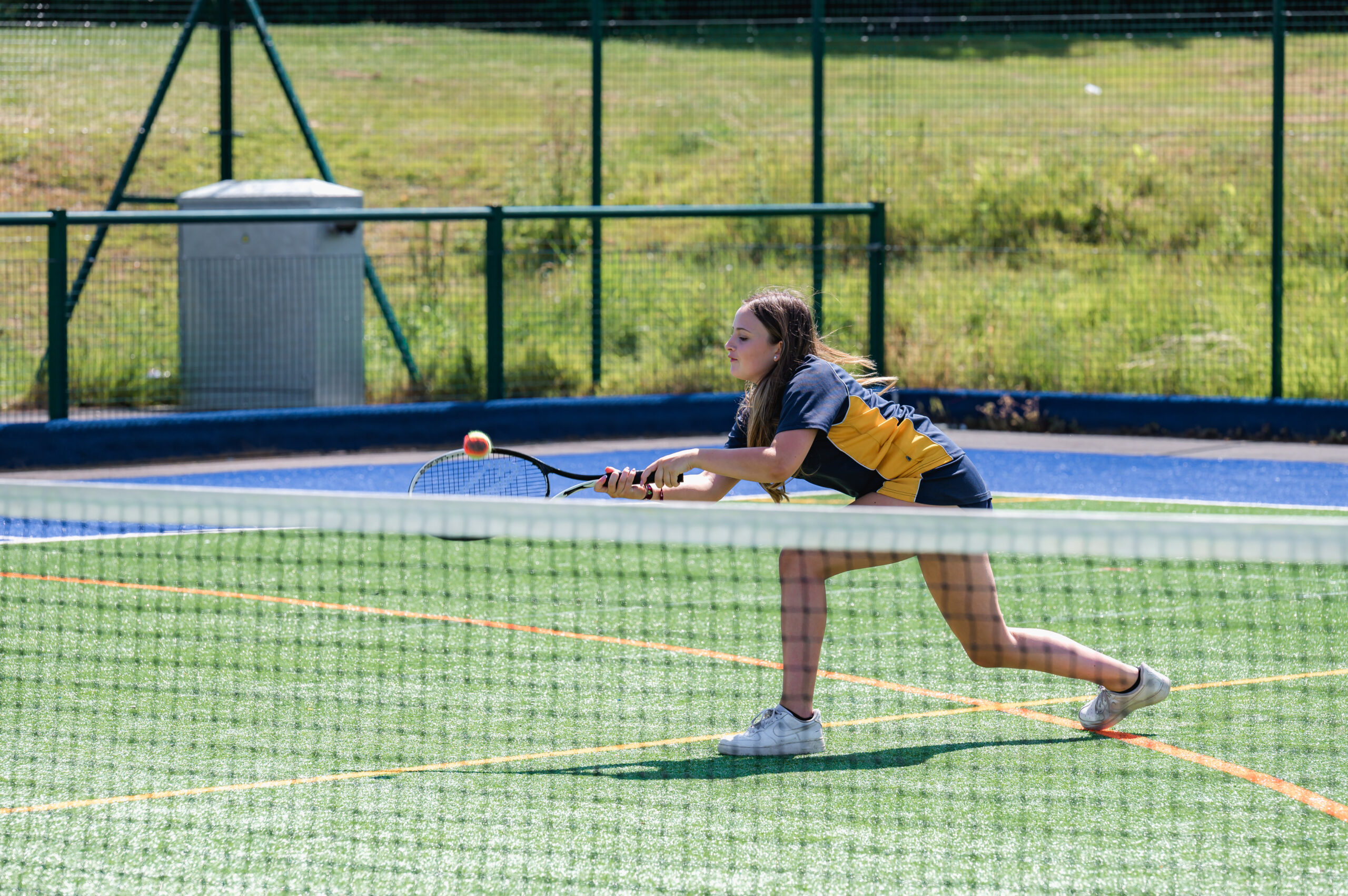 A pupil, wearing a blue and yellow P.E kit, it playing tennis on an outdoor tennis court. She is reaching over to hit the ball with the racket in her right hand.