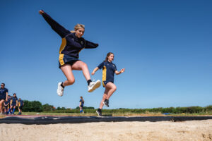 Two girls, dressed in yellow and blue P.E kits, are jumping into a large container of sand.