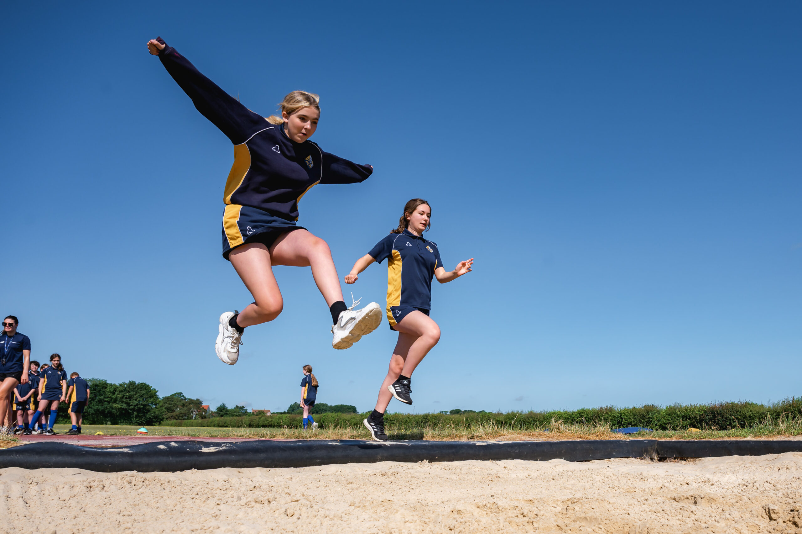 Two girls, dressed in yellow and blue P.E kits, are jumping into a large container of sand.