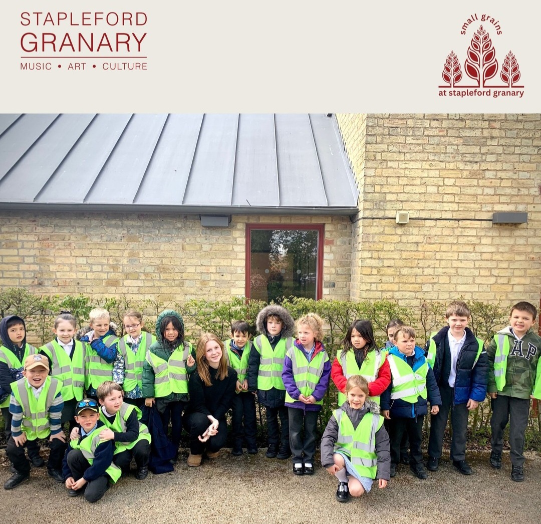 A group of young pupils are standing in front of a building smiling at the camera. They are all wearing high-vis jackets.