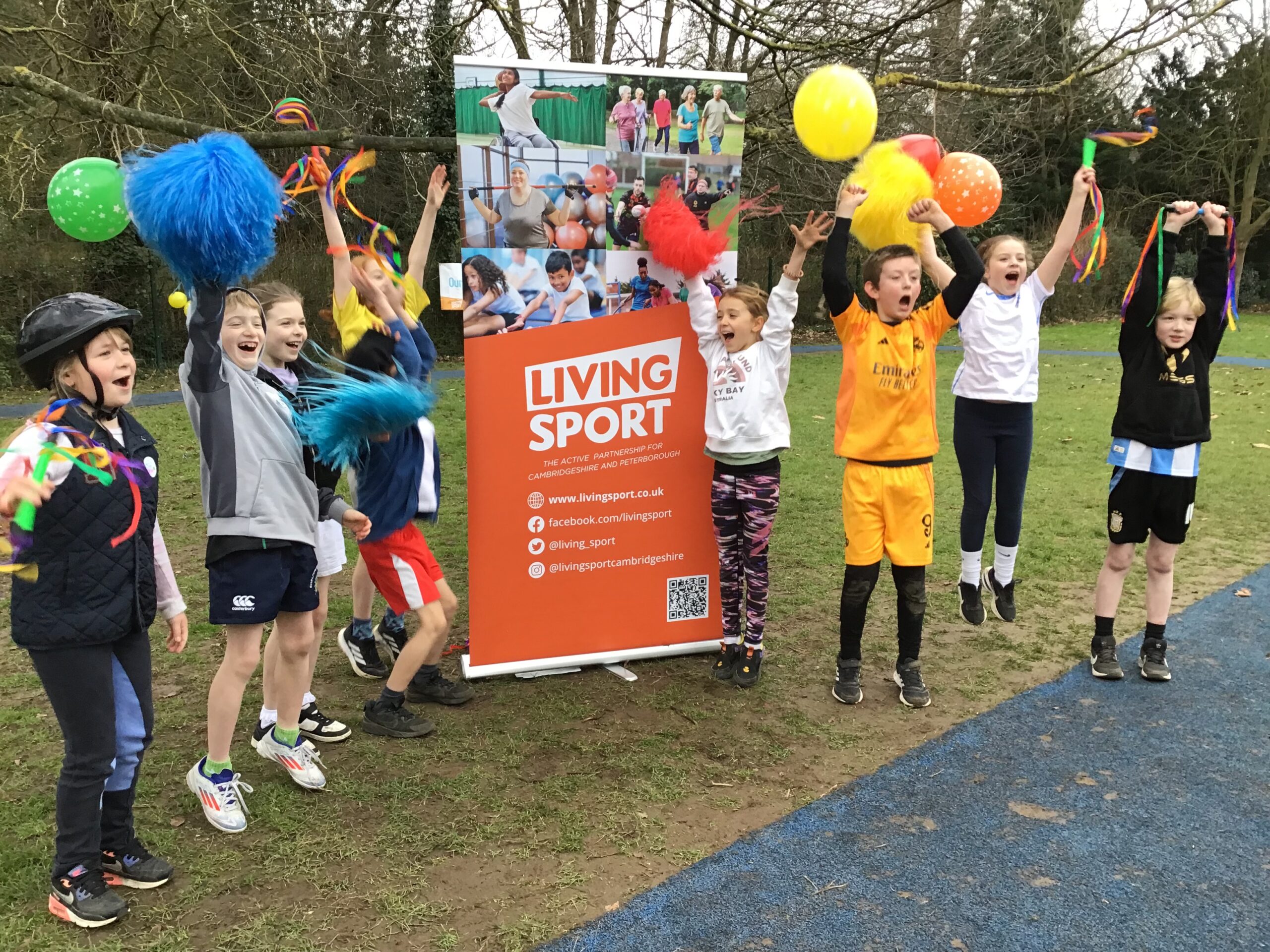 A group of pupils are standing next a sign that reads 'living sport'. They are wearing colourful outfits and cheering to the camera.
