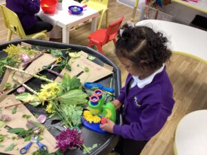 A pupil is sitting at a table and is putting a flower arrangement together. They are wearing a purple jumper with a white collar.