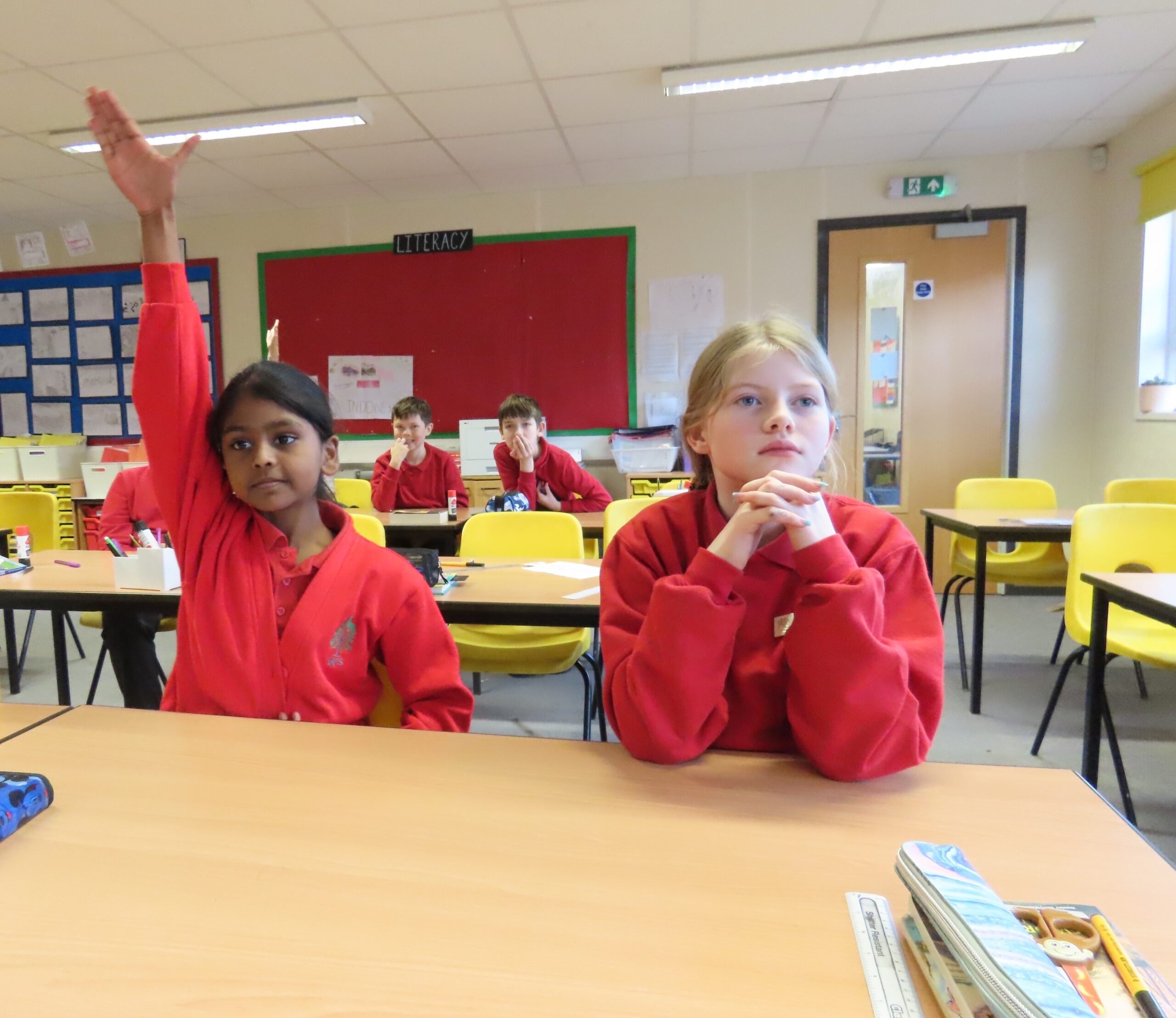 Two children sitting at a desk, one has their hand up