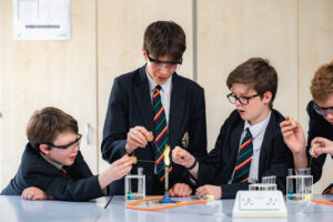 Four pupils are sitting at a desk. They are all wearing a black blazer with a stripey tie. They are wearing safety glassed and carrying out a science experiment.