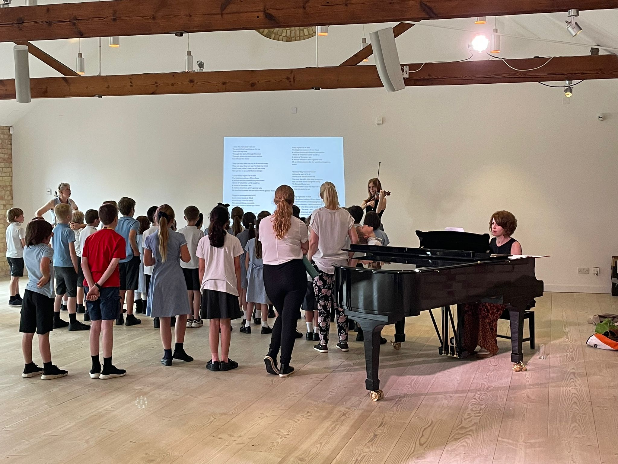 A group of pupils are standing next to a piano which is placed infront of a large projector screen.