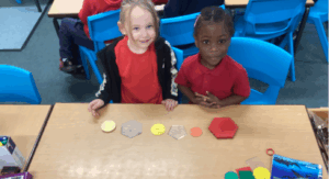 Two pupils are sitting at a desk. They have colourful tokens laid out in front of them. They are smiling to the camera.