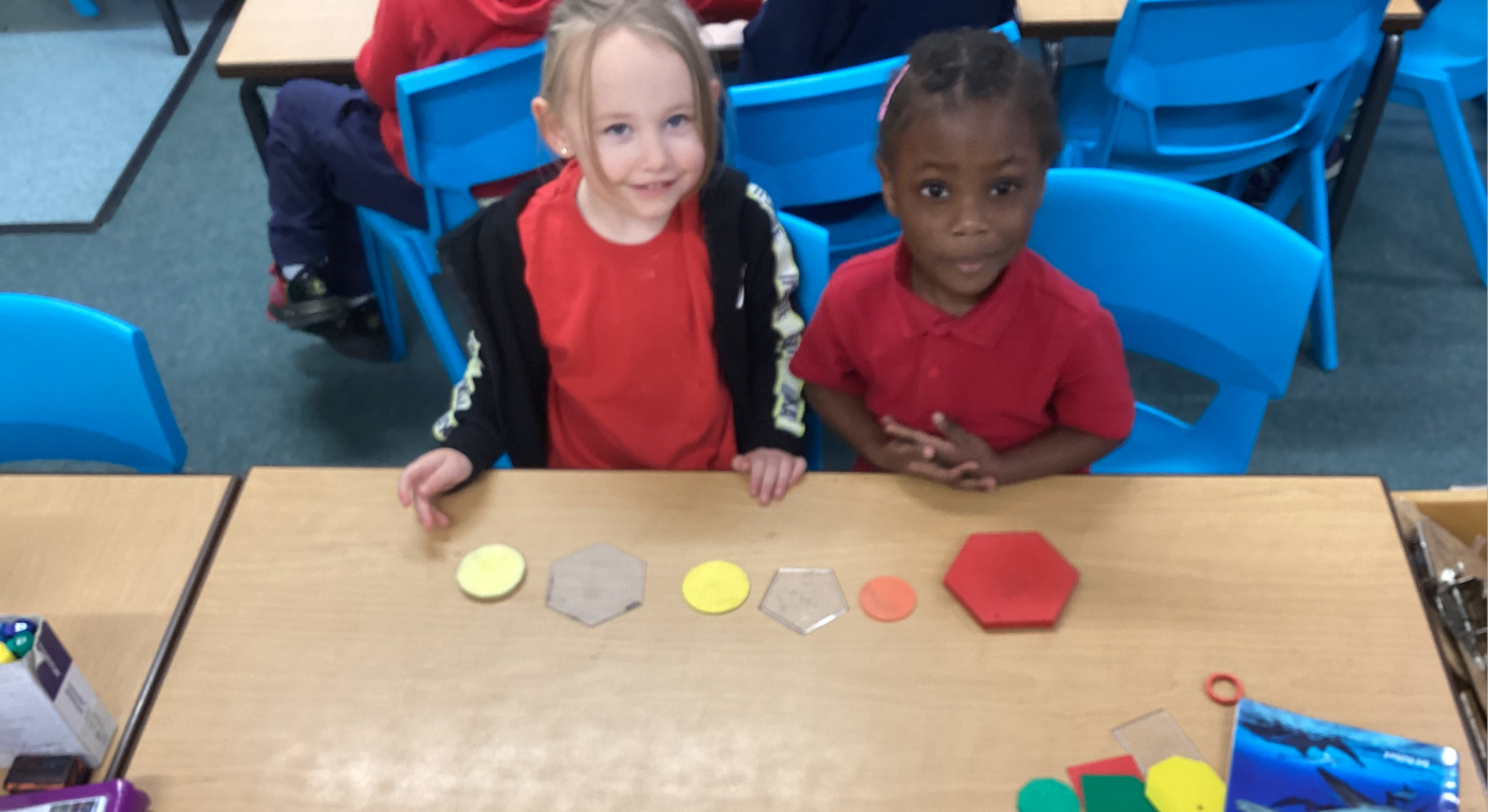 Two pupils are sitting at a desk. They have colourful tokens laid out in front of them. They are smiling to the camera.