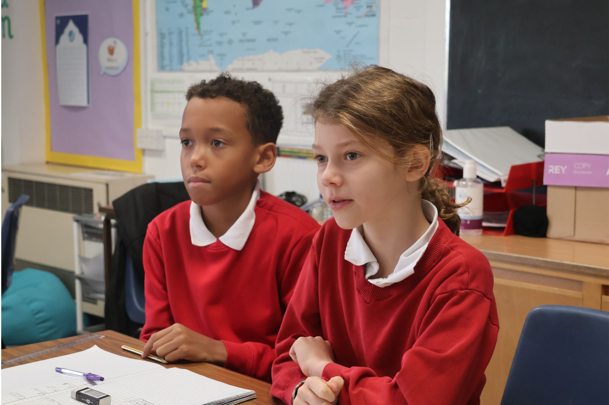 Two pupils are sitting at a desk. They are wearing red jumpers with a white collar sticking out.
