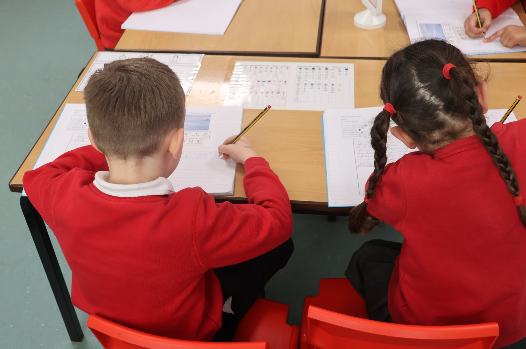 Two pupils are sitting at a desk, writing on a piece of paper. They are wearing red jumpers.
