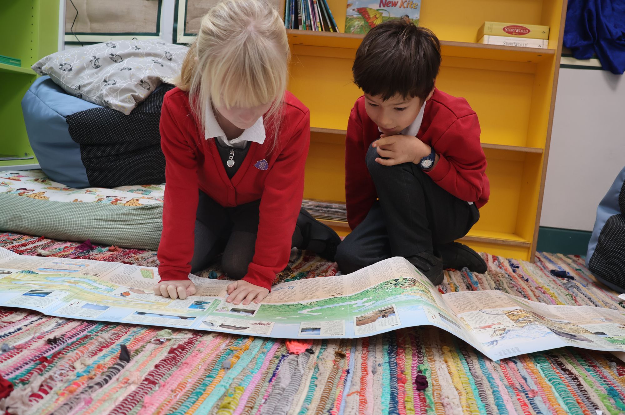 Two pupils are kneeling on the ground. They are reading a large book.
