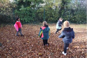 Four pupils are playing outside in fallen leaves next to a large tree. They are all wearing thick coats and hats.