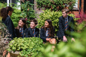 Five pupils are sitting outside near a large green bush. They are all wearing a black jacket and stripey tie. They are facing each other and talking.