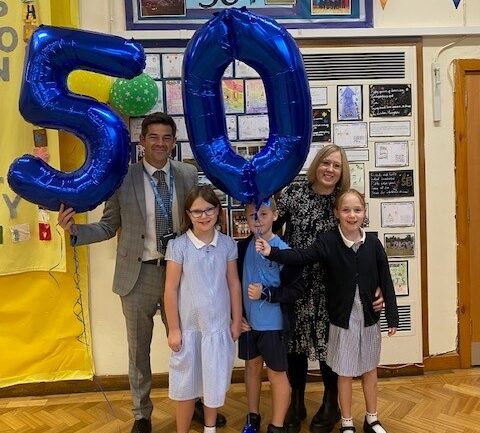 Two adults stand with three pupils and a two large balloons which display the number '50'