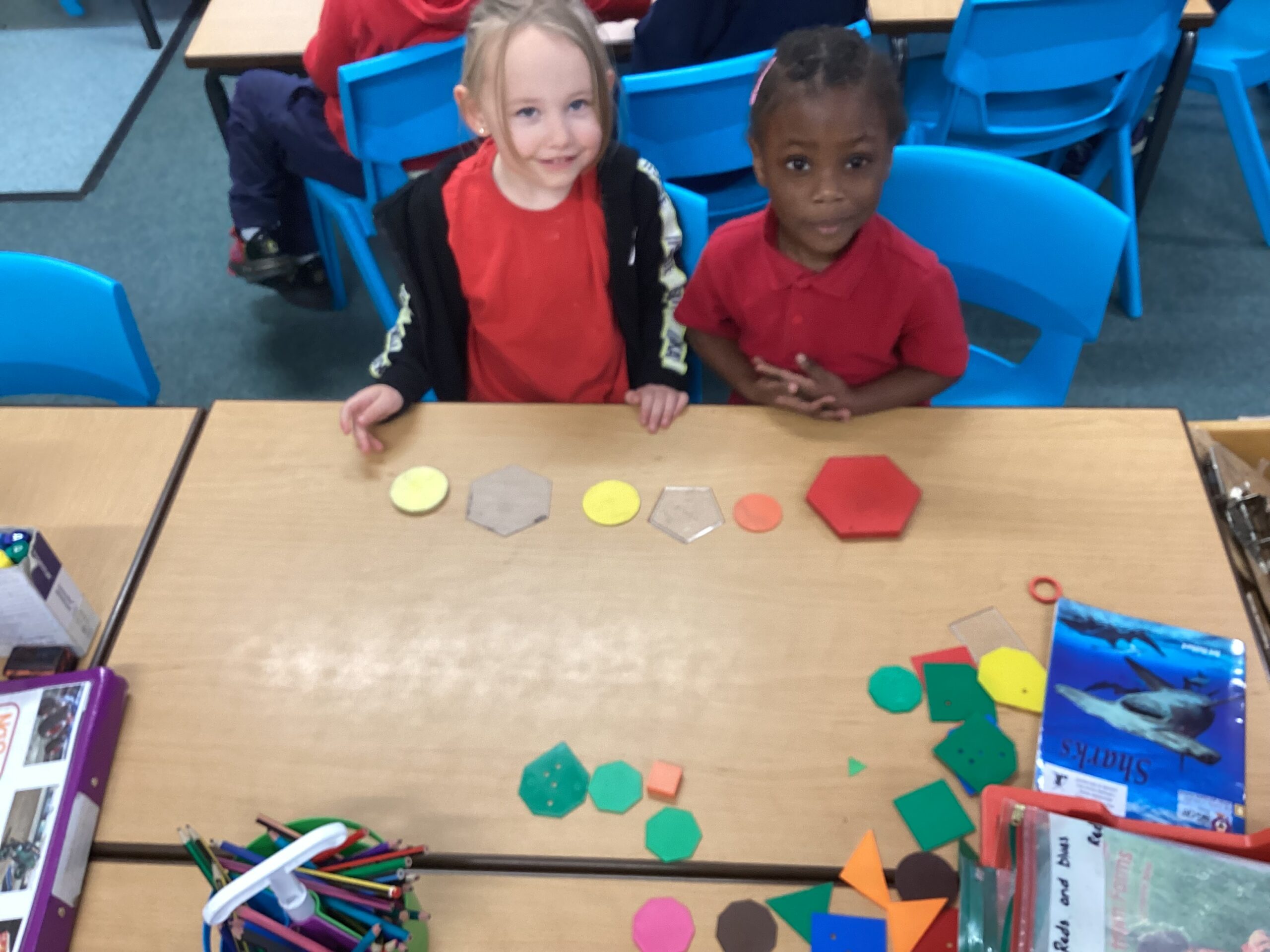 Two children, wearing red polos sitting at a desk that has random shapes in different colours