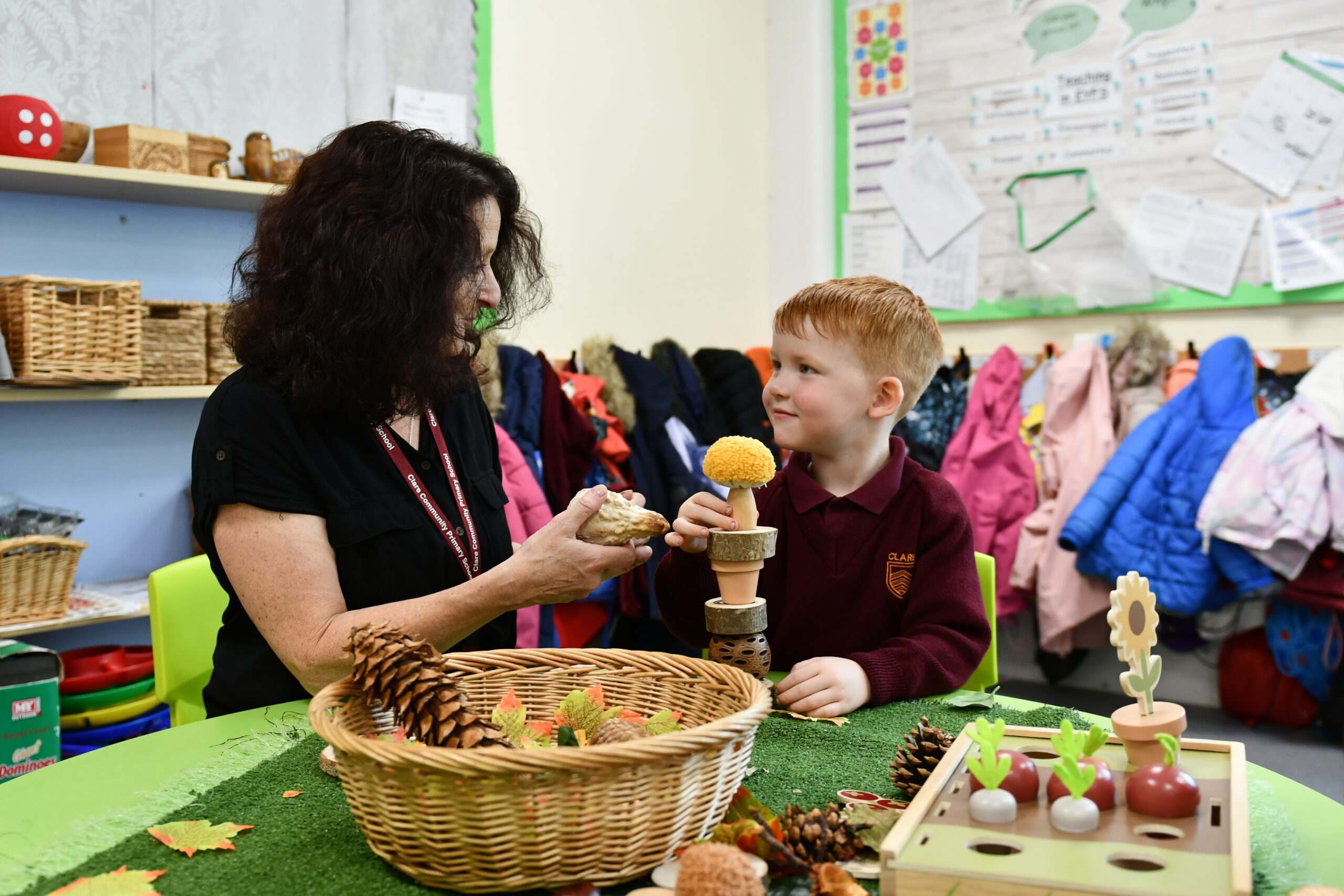 A teacher is sitting at a desk with a young white boy with ginger hair. They have a basket full of pine cones in front of them which they are stacking on top of eachother on the table.