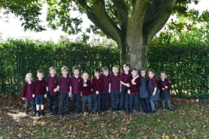 A large group of pupils are standing underneath a tree. They are all wearing school uniform which is a burgundy jumper with black shorts and trousers.