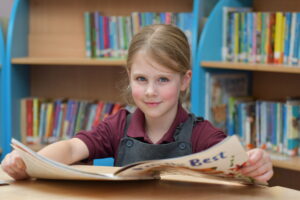 A young white girl with blonde hair is smiling at the camera whilst sitting at a desk reading a book.