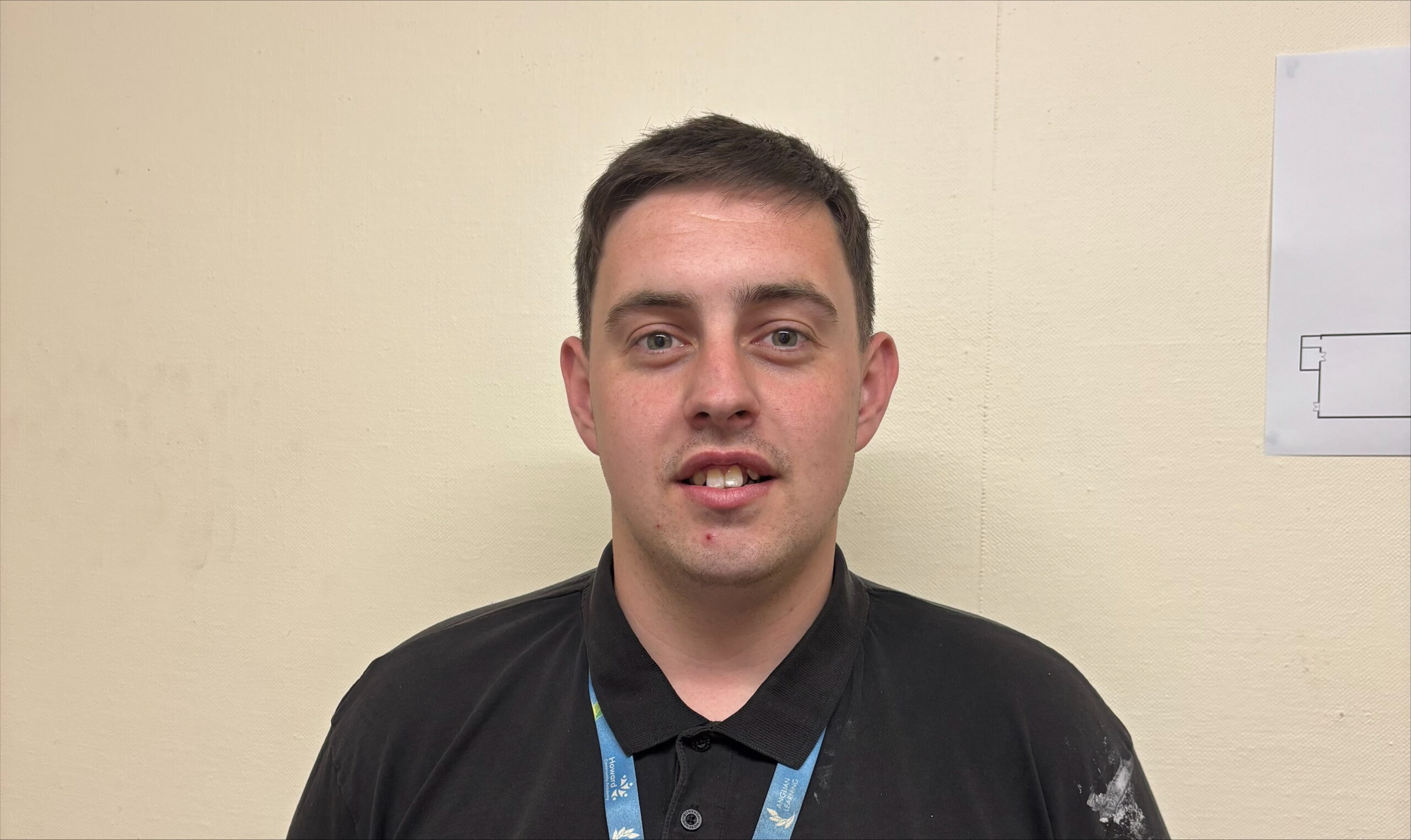 Tom Chaplin_Site Officer at Howard Community Academy A man is standing in front of the camera smiling. He is wearing a blue polo neck and blue lanyard.