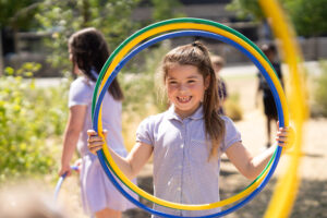 A pupil wearing a purple checkered dress is holding up three hula hoops to her head and smiling to the camera within the middle circle.