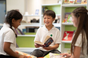 A young pupil in a white polo top is reading from a book to two other pupils who are also wearing white polo tops.