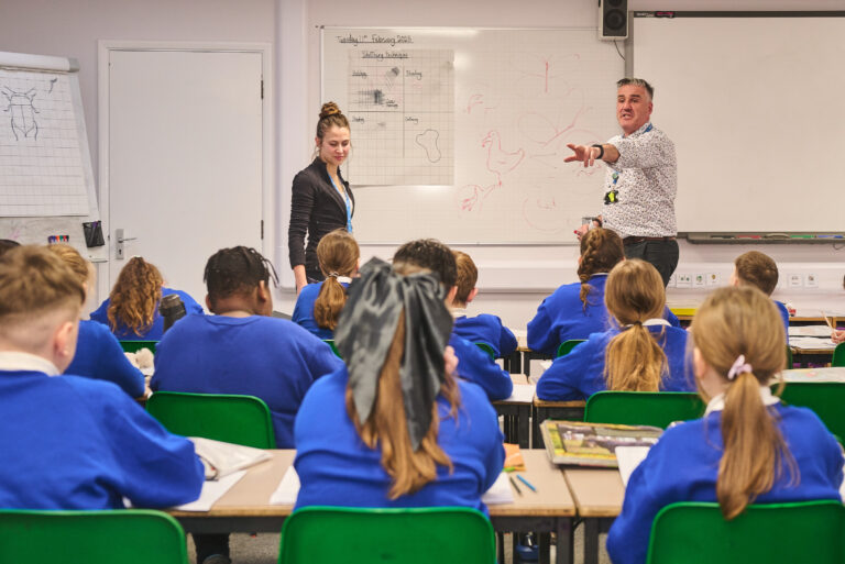 Two teachers are standing at the front of a classroom in front of a whiteboard. One teacher is pointing at a pupil, facing towards them, with their hand up.