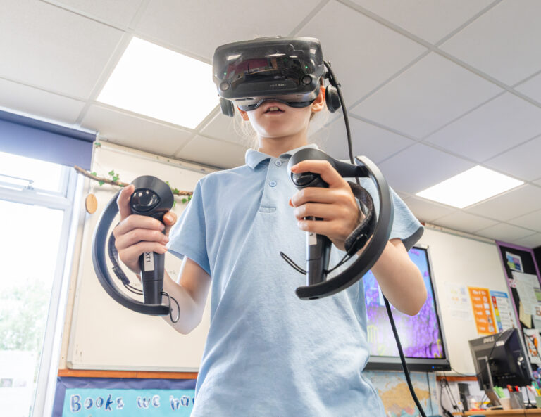 A young pupils wearing a blue polo is wearing a black VR headset and holding two black controllers in his hands.