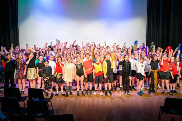A large group of pupils are standing on a stage. They are wearing a mixture of bright colours. They are all cheering with their hands up in the air and smiling at the camera.