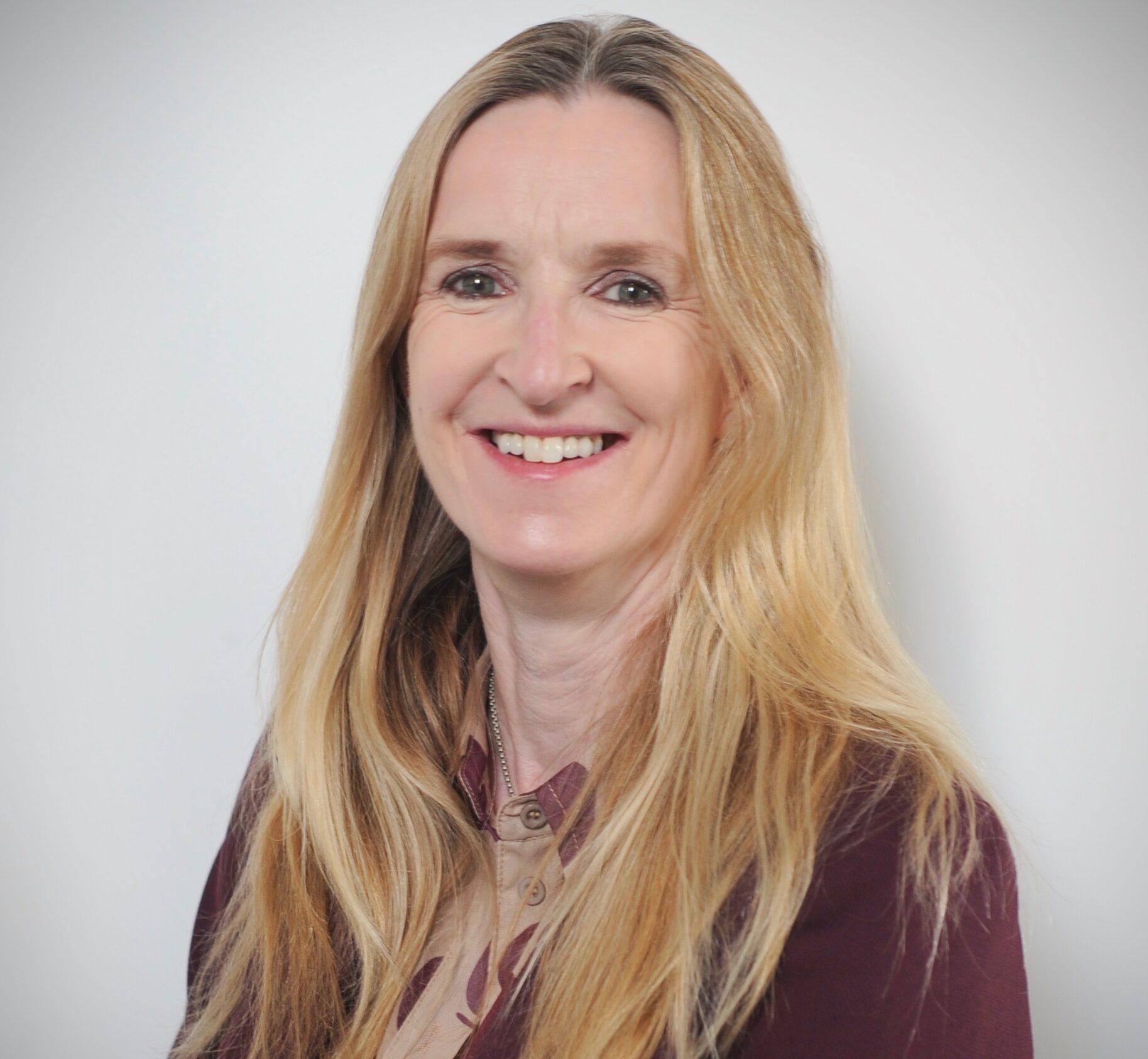 A white woman with long blonde hair who is wearing a purple cardigan and and pink top is smiling at the camera.