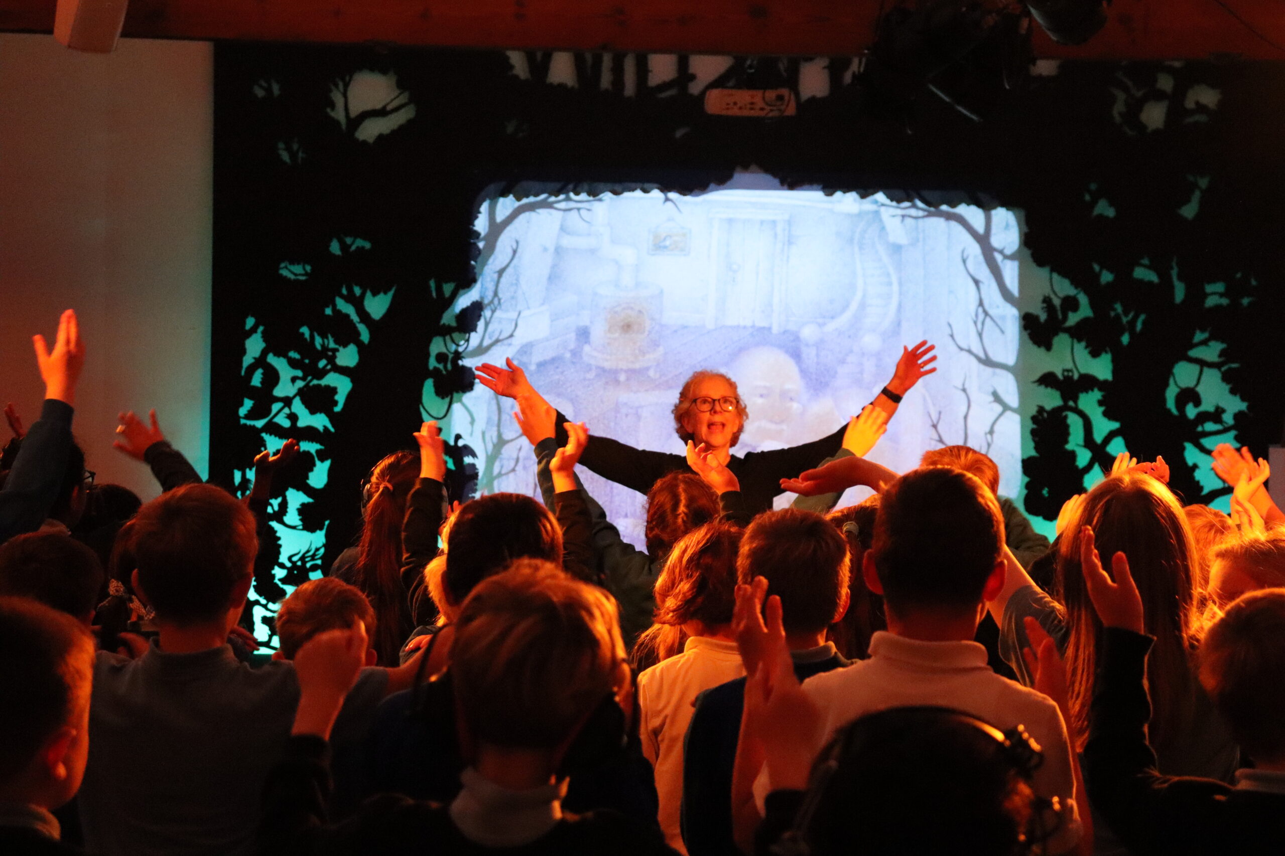 IMG_7580 A woman with short, blonde hair is standing with her arms extended facing a crowd of pupils. The pupils, with their backs towards the camera, are also standing with the arms extended, copying the woman.