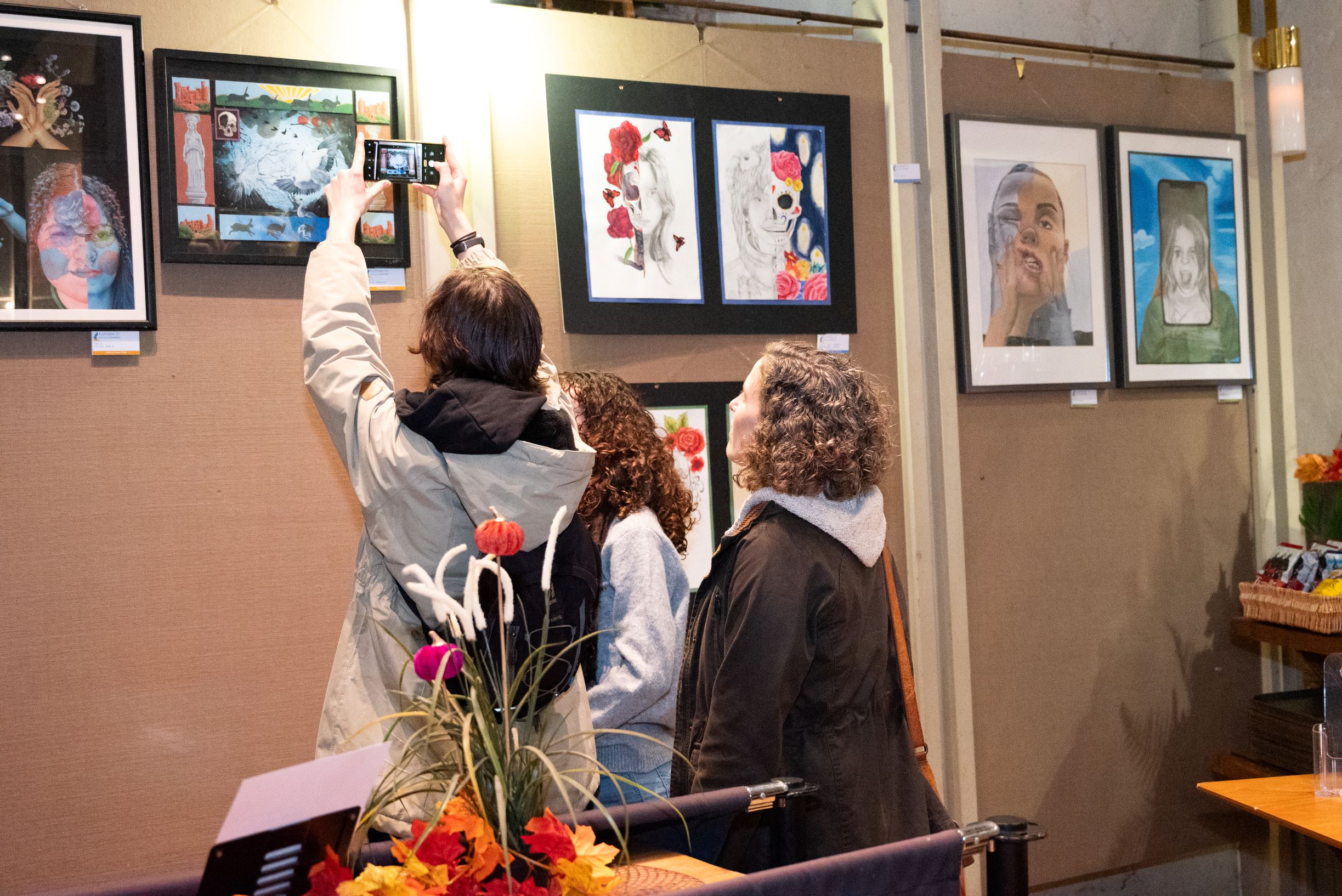 A group of people are viewing artworks hanging on a wall. One person is taking a photograph of the artwork.