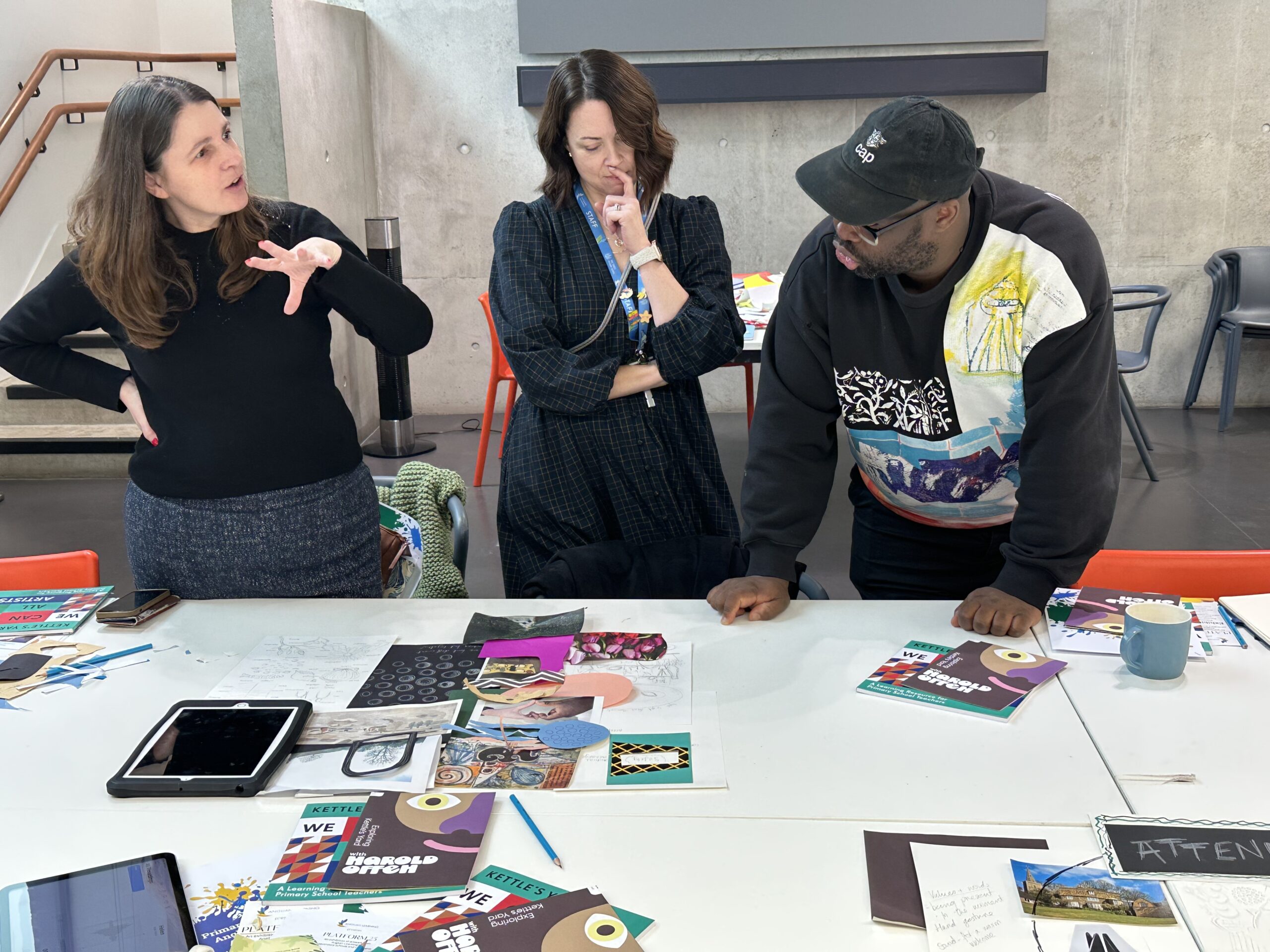 Three people are leaning over a desk. Two women, one wearing a black top and blue skirt, another wearing a patterned dress, are speaking with a man, wearing a black jumper and cap.