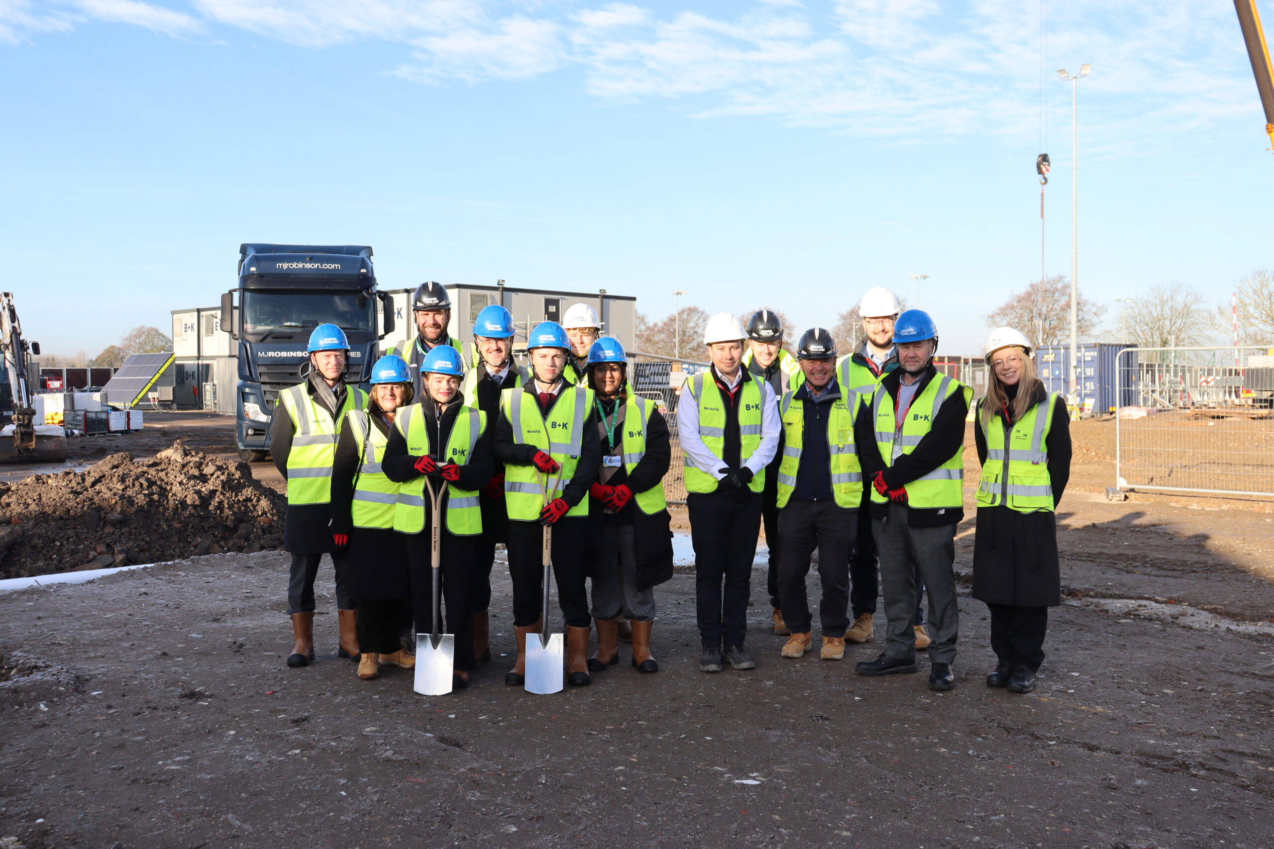 A group of people are standing on a construction site. Two pupils are holding spades. All are wearing yellow reflective jackets and blue and white hard helmets.